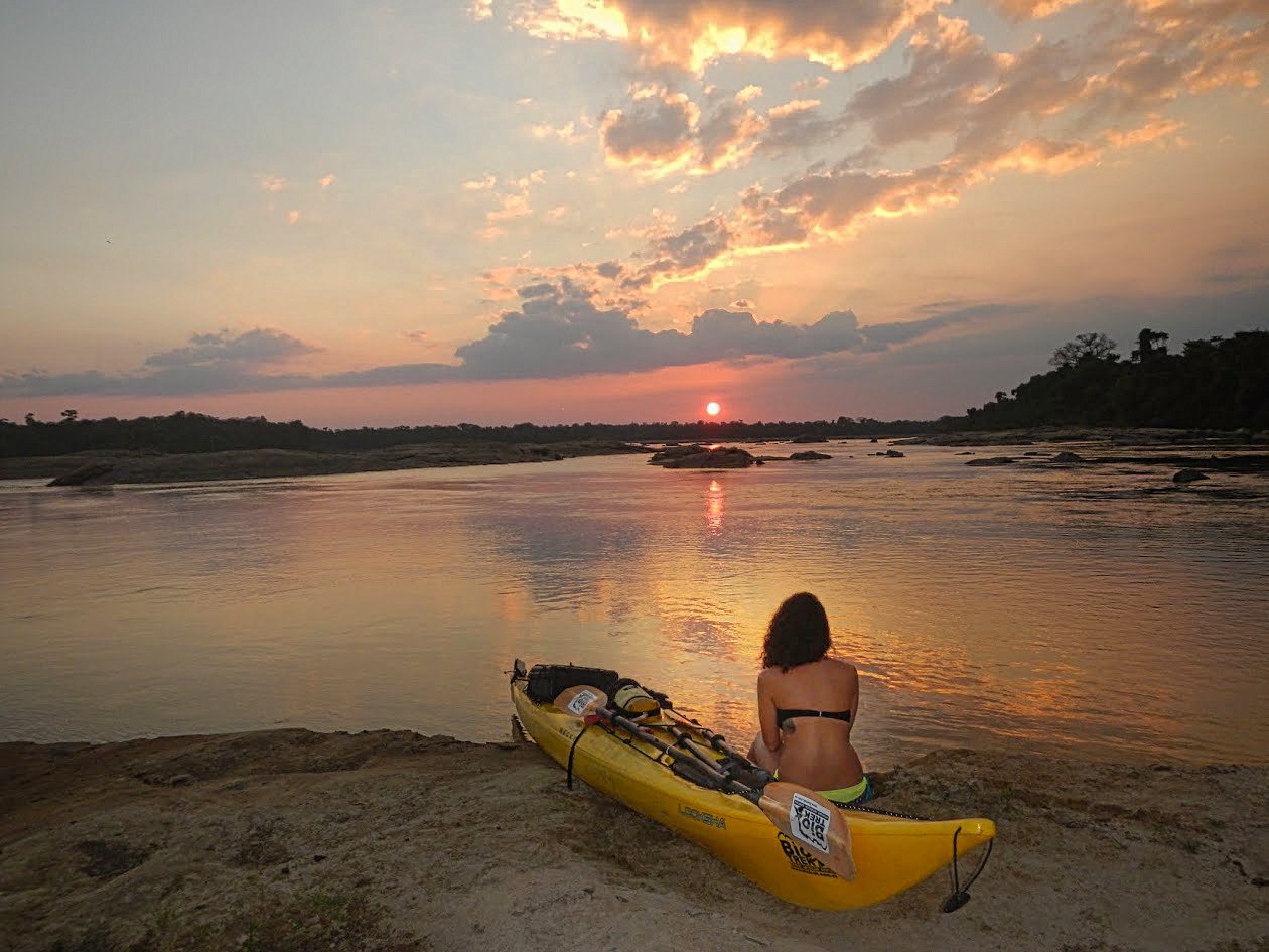 Expedicionaria sentada junto a su kayak frente al atardecer en una playa del río Caura