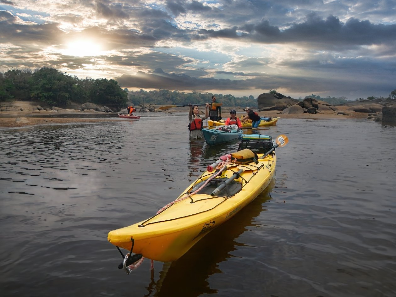 Grupo de kayakistas con sus embarcaciones en el río Caura bajo un cielo de nubes al atardecer
